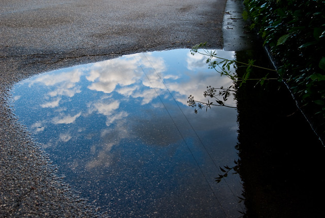 image of a pot hole puddle on the sideof the road with the reflection showing a partially clowdy blue sky