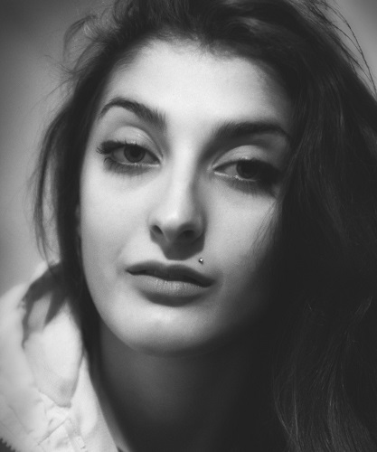 Black and white image of a woman looking at the reader with long messy hair in a blank room